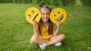 A smiling girl holds up one frowning face sign and one happy face sign.