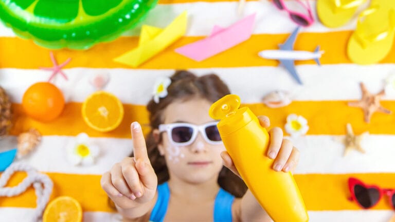 Young girl laying on beach towel raising sunscreen into the camera