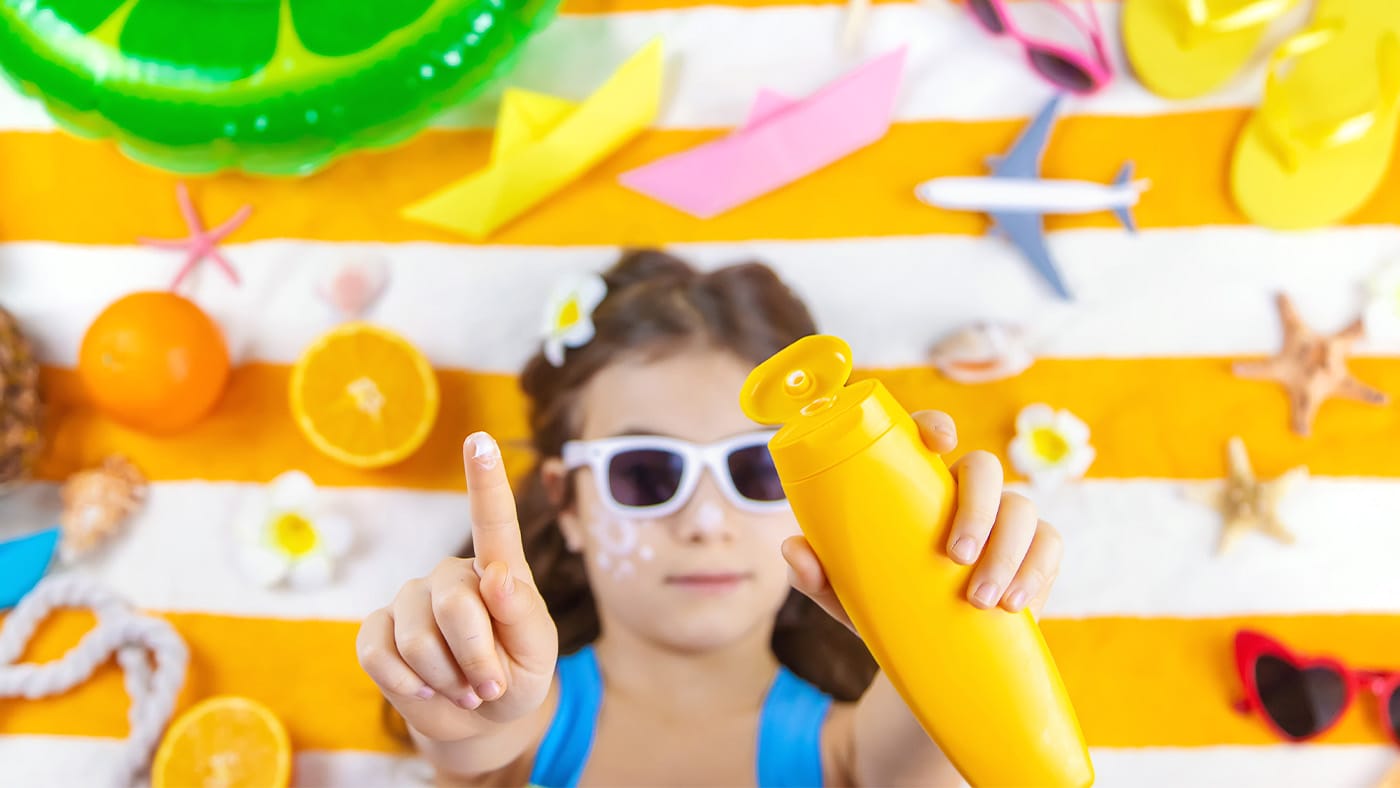 Young girl laying on beach towel raising sunscreen into the camera
