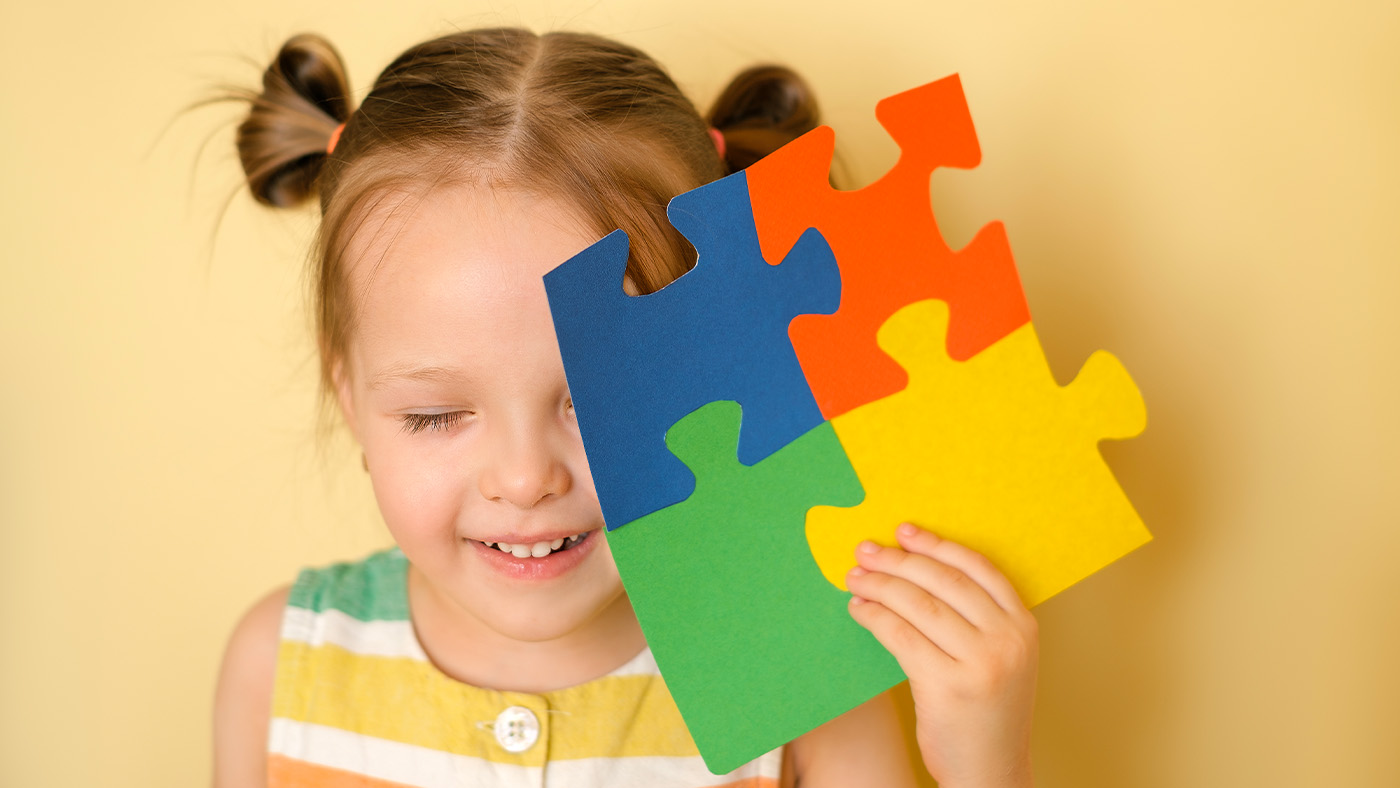 Little girl with autism holding up puzzle pieces in colors representing Autism Awareness Week
